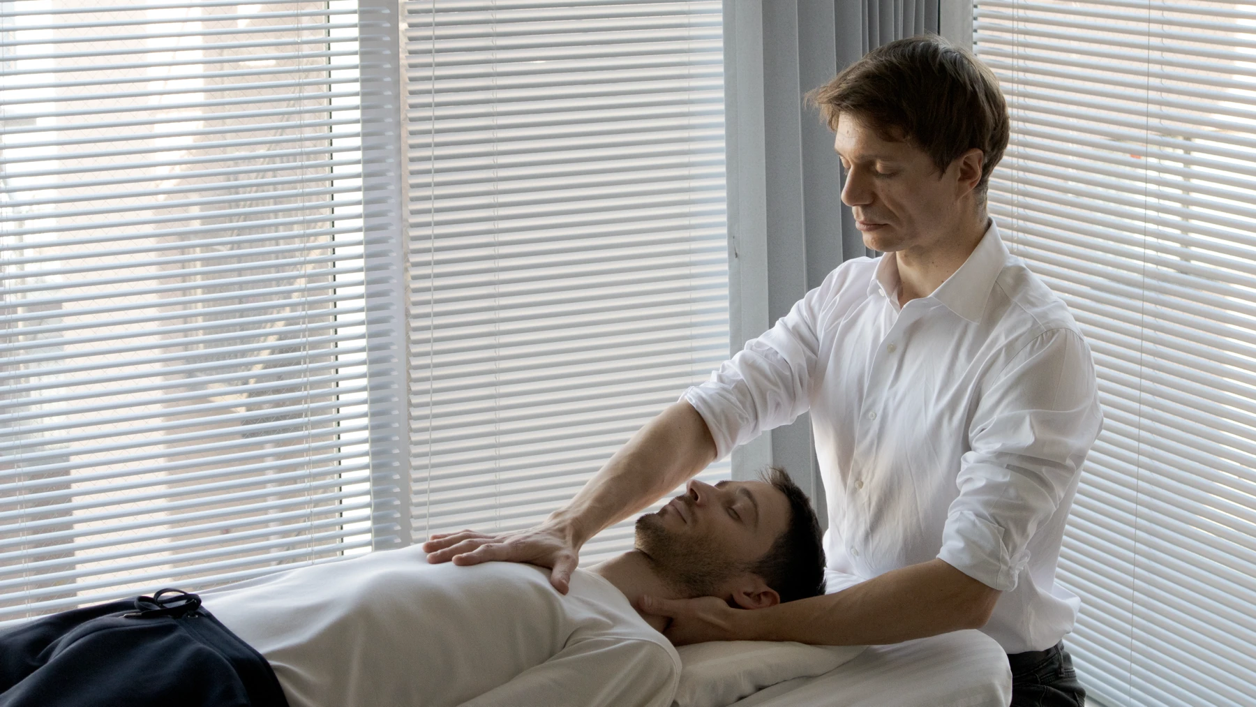 David Brisson D.O. stands at the head of the treatment table in bright window light, one hand under the neck and the other resting still on the chest of a patient lying face up with their eyes closed.